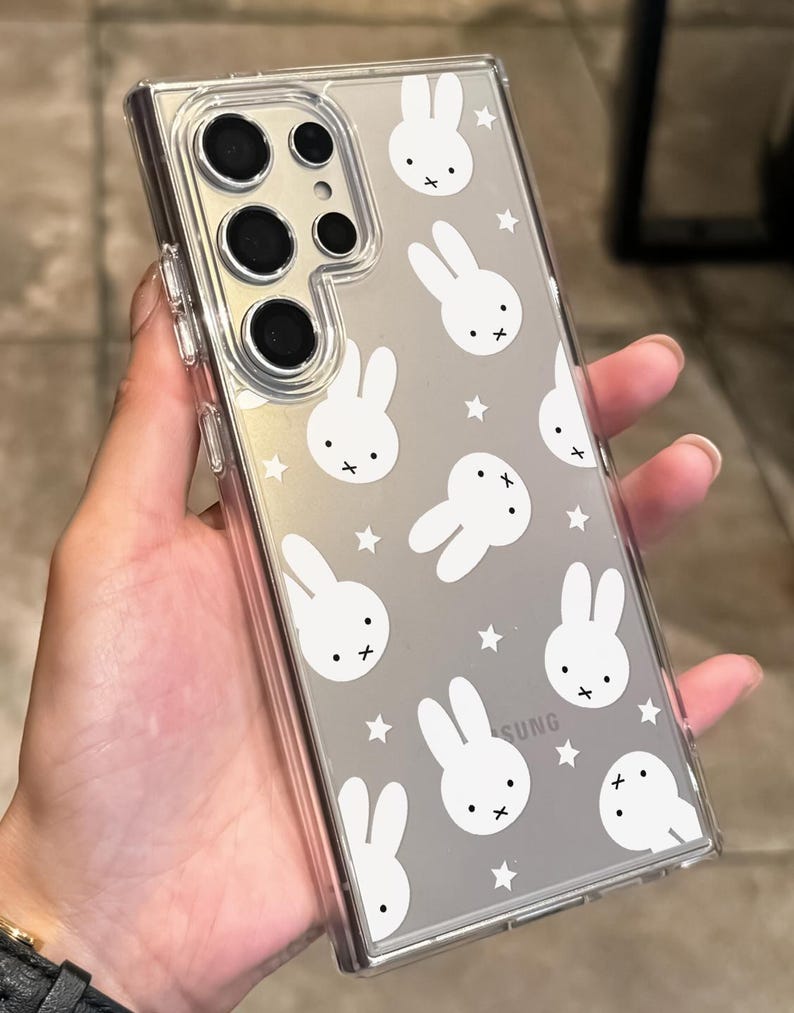 May include: Clear phone case with a white bunny and star pattern. The case has a clear border and covers the back of the phone. The phone has multiple camera lenses.