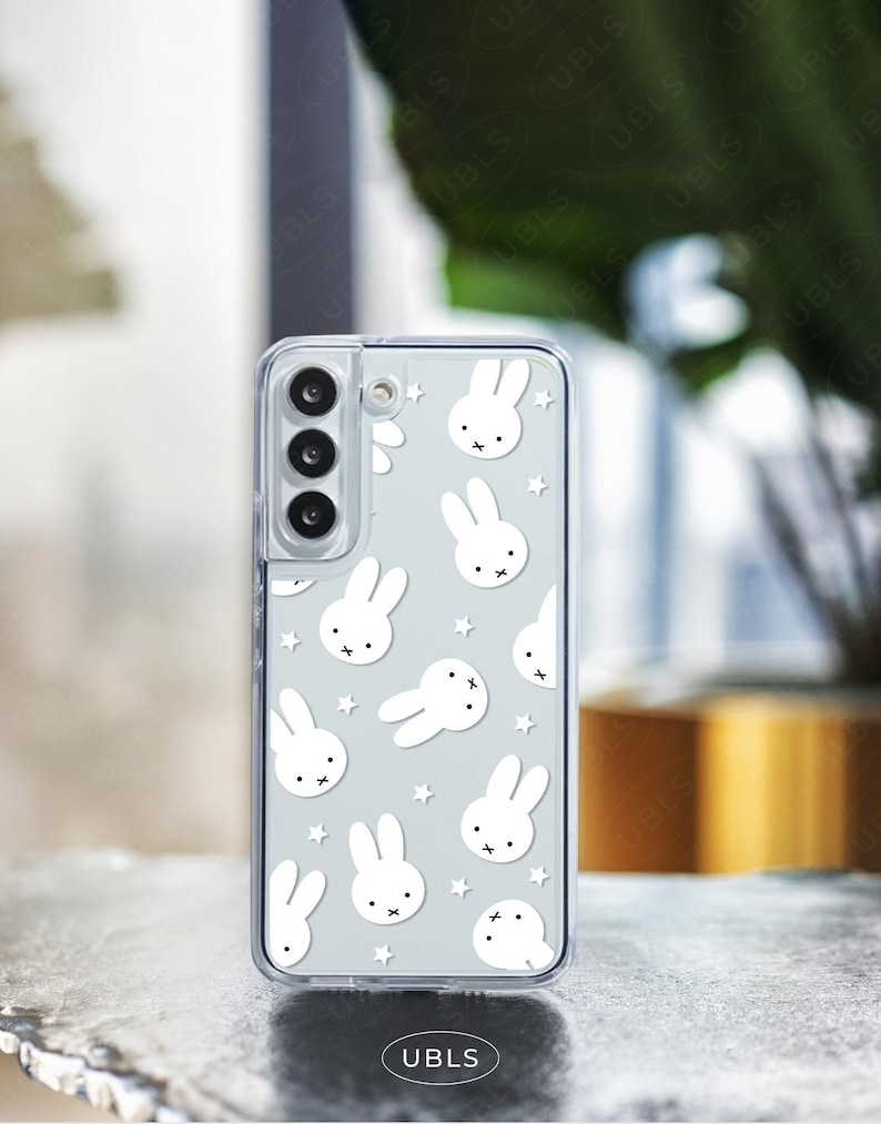 May include: A clear phone case featuring a pattern of white bunny faces and stars on a light gray background. The phone case has a camera cutout and the text "UBLS" at the bottom.
