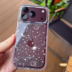 May include: A clear smartphone case featuring a white celestial print of stars, planets, and crescent moons. The case is fitted over a dark maroon phone with a triple-lens camera system and a visible Apple logo on the back. The text UBLS is printed.