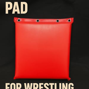 May include: Bright red turnbuckle pad with reinforced grommets, designed for wrestling and boxing. The pad is rectangular and features the text "TURNBUCKLE PAD" at the top and "FOR WRESTLING & BOXING" at the bottom, all in a bold, sans-serif font.