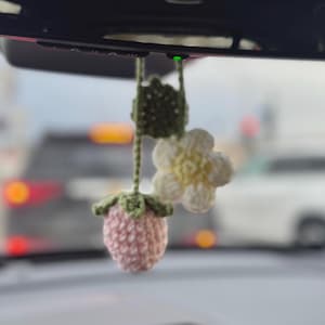 May include: A handmade crochet car charm featuring a pink strawberry, a white and yellow flower, and a green leaf. The charm is suspended from a string, adding a touch of whimsy to any vehicle's interior.