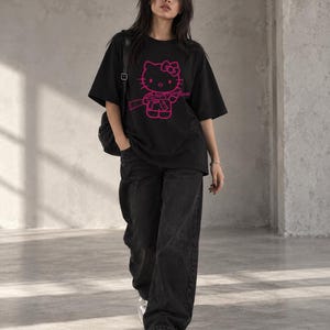 Puede incluir: Una camiseta negra de manga corta con un gráfico rosa brillante de un personaje de gato de dibujos animados que sostiene un rifle. La prenda se combina con pantalones vaqueros negros holgados y zapatillas blancas, creando un conjunto casual.
