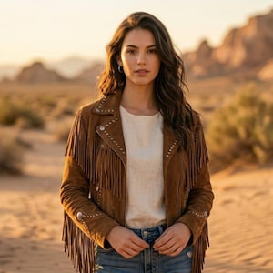 May include: A woman wearing a brown suede jacket with fringe and silver studs, paired with a white top and blue jeans. The jacket is the focal point, set against a desert backdrop with golden sunlight.