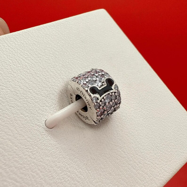 Puede incluir: Un charm de plata con un dise&ntilde;o de cabeza de Mickey Mouse. El charm est&aacute; incrustado con peque&ntilde;os cristales transparentes y presenta una cabeza de Mickey Mouse negra. El charm est&aacute; sobre un fondo blanco.