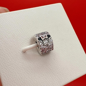 Puede incluir: Un charm de plata con un dise&ntilde;o de Minnie Mouse. El charm presenta un lazo negro y rojo, orejas negras y est&aacute; incrustado con cristales transparentes y rosas. El charm est&aacute; sobre un palito blanco, sobre un fondo blanco.