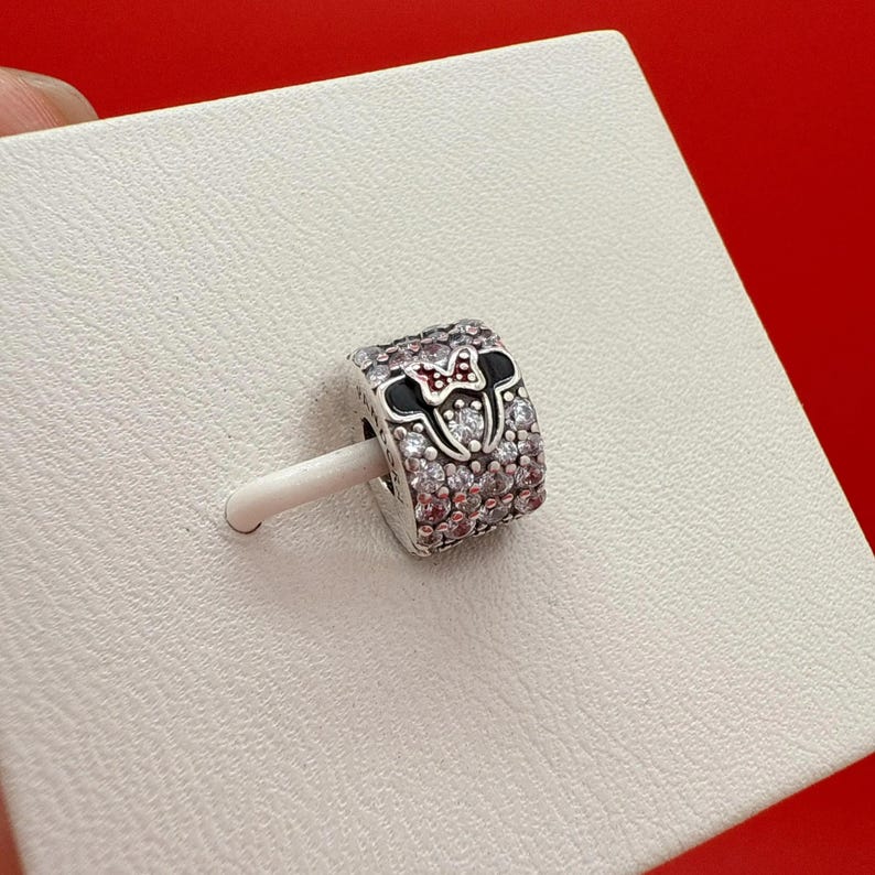 Puede incluir: Un charm de plata con un dise&ntilde;o de Minnie Mouse. El charm presenta un lazo negro y rojo, orejas negras y est&aacute; incrustado con cristales transparentes y rosas. El charm est&aacute; sobre un palito blanco, sobre un fondo blanco.