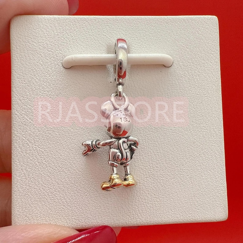 May include: A silver charm featuring a Mickey Mouse figure with gold-colored shoes and accents. The charm is suspended from a silver loop and is attached to a white card. The text "100" is visible on the charm.