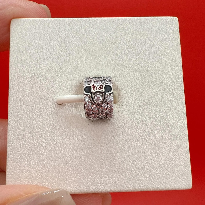 Puede incluir: Un charm de plata con un dise&ntilde;o de lazo de Minnie Mouse. El charm presenta una banda negra con un lazo rojo y cristales transparentes. El charm est&aacute; sobre un fondo blanco.