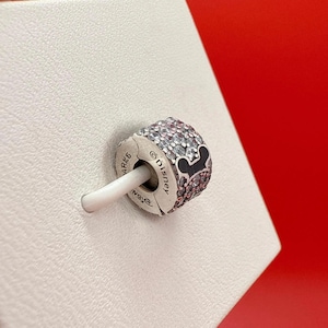 Puede incluir: Un charm de plata con un dise&ntilde;o de Mickey Mouse. El charm est&aacute; incrustado con cristales transparentes y rosas. La silueta de Mickey Mouse es negra. El charm est&aacute; marcado con el logotipo de Disney y otro texto.