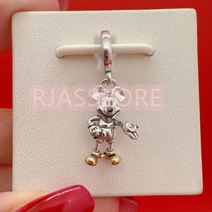 May include: A silver Mickey Mouse charm with gold-colored shoes and a small clear gem on his right hand. The charm is suspended from a silver loop and is displayed on a white card. The text "RJASS STORE" is visible.