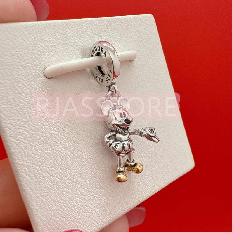 May include: A silver and gold Mickey Mouse charm with a pointing gesture. The charm is attached to a silver ring with the word "Pandora" engraved on it. The charm is displayed on a white surface against a red background.
