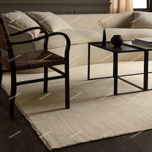 Puede incluir: Alfombra beige tejida con borde deshilachado, colocada sobre un suelo de madera oscura. Una silla de madera marrón oscuro con asiento tejido y una mesa de centro de metal negro están sobre la alfombra. Un sofá crema con cojines está en el fondo.