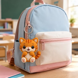 Puede incluir: Una mochila de color azul claro y rosa con un bolsillo frontal y un llavero de metal plateado que presenta un pequeño gato atigrado naranja de ganchillo con detalles blancos, ojos negros y almohadillas rosas en las patas.