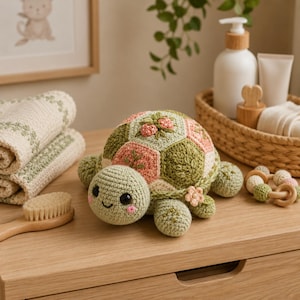 Op de afbeelding: Een gehaakt schildpadspeeltje met een schild bestaande uit zeshoekige vlakken in saliegroen, crème en oudroze. Het schild is versierd met kleine geborduurde bloemmotieven en een gehaakt bloemetje. De schildpad heeft zwarte kraaloogjes.