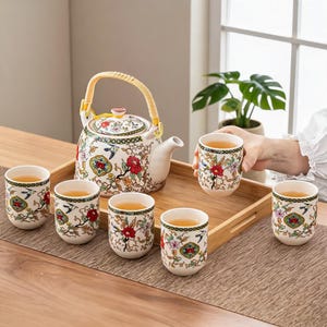 Puede incluir: Un juego de té de cerámica que incluye una tetera con asa de bambú curvada y cinco tazas a juego. La porcelana blanca está decorada con un colorido patrón floral con motivos rojos, rosas y verdes, junto con pequeños pájaros azules y bordes.