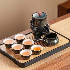 May include: A black ceramic tea set with six small cups filled with tea, a teapot, and a serving pot on a bamboo tray. The teapot has a blue lid with a red knob and gold accents.