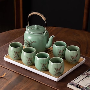 May include: A celadon green tea set comprising a teapot with a woven handle and six matching cups. Each piece is decorated with a delicate floral design in red and brown. The set sits on a white tray with a bamboo border, ideal for serving tea.