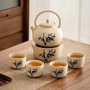 Peut inclure: Service à thé en céramique beige comprenant une théière, un chauffe-plat et quatre tasses. Chaque pièce est ornée d'un motif de bambou bleu. Les tasses sont remplies de thé, posées sur une table en bois.