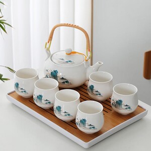 Op de afbeelding: Een witte keramische theeset met een theepot en zes bijpassende kopjes, allemaal versierd met een blauw-groen landschapsontwerp. De theepot heeft een geweven handvat en staat op een bamboe dienblad. De kopjes zijn ongeveer 7.6 cm hoog.