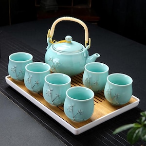 Puede incluir: Juego de té de cerámica azul claro con una tetera y seis tazas a juego. La tetera tiene un asa trenzada y un diseño floral. Las tazas y la tetera están dispuestas sobre una bandeja de bambú con borde blanco. Para el té.