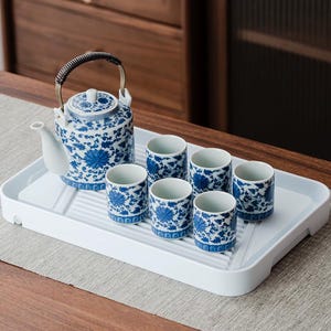 Puede incluir: Un juego de té de porcelana azul y blanca con una tetera y seis tazas a juego. La tetera tiene un asa de metal y una tapa. Las tazas y la tetera están decoradas con un estampado floral. El juego está sobre una bandeja blanca.