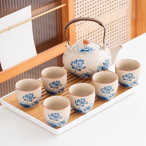 Puede incluir: Juego de té de cerámica crema con motivos florales azules, que incluye una tetera con asa trenzada y seis tazas a juego. El juego se presenta sobre una bandeja de bambú.