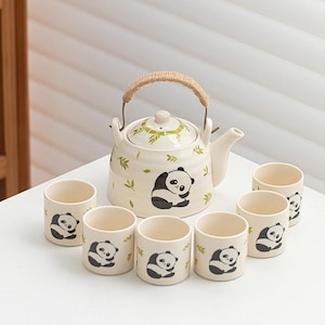 Puede incluir: Un juego de té color crema con una tetera y seis tazas, cada una adornada con un diseño de oso panda en blanco y negro. La tetera tiene un asa tejida y una tapa con detalles de hojas verdes. Las tazas están dispuestas alrededor de la tetera.