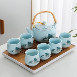 Op de afbeelding: Een lichtblauwe keramische theeset met een theepot en zes bijpassende kopjes, elk versierd met een landschapsschilderij. De theepot heeft een geweven handvat. De set wordt gepresenteerd op een bamboe dienblad.