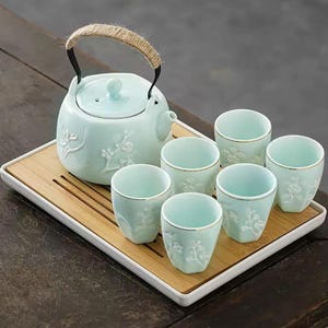 Op de afbeelding: Een lichtblauwe keramische theeset met een theepot, zes kopjes en een serveerblad. De theepot heeft een geweven handvat en bloemreliëfdetails. De kopjes hebben gouden randen en bloemmotieven. De set staat op een houten dienblad met een witte rand.