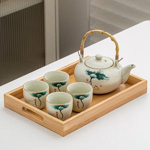 Puede incluir: Juego de té de cerámica crema con tetera y cuatro tazas, presentado en una bandeja de bambú. La tetera tiene un asa de bambú y un diseño floral verde y azul. Las tazas presentan un diseño similar.