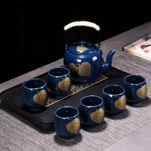 Op de afbeelding: Een blauwe keramische theeset met een theepot en zes kopjes. Elk stuk heeft een gouden bladeren ontwerp. De theepot heeft een witte handgreep. De set is gerangschikt op een zwarte dienblad, ideaal voor het serveren van thee.