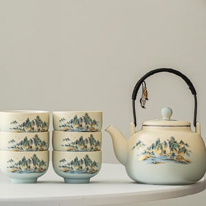 Puede incluir: Un juego de té con una tetera y seis tazas a juego. La tetera tiene un asa negra y una tapa azul claro. Las tazas y la tetera están decoradas con un diseño de paisaje en tonos azules, verdes y dorados.