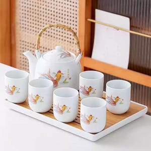May include: A white ceramic tea set with a teapot and six cups, each adorned with a colorful fish design. The teapot has a bamboo handle. The set sits on a rectangular wooden tray, perfect for serving tea.
