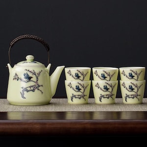 Könnte beinhalten: Hellgelbes Keramik-Teeservice mit einer Teekanne mit geflochtenem Griff und sechs passenden Tassen. Jedes Stück ist mit einem blauen Vogel auf einem blühenden Ast verziert. Das Set wird auf einer neutralen Oberfläche präsentiert.