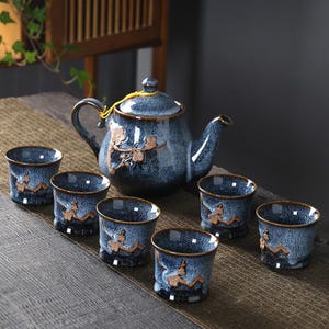 Könnte beinhalten: Ein blaues Keramik-Teeservice mit einer Teekanne und sechs passenden Tassen. Die Teekanne und die Tassen haben eine gesprenkelte Oberfläche und ein braunes Blumendesign. Das Set ist auf einer Holzoberfläche angeordnet.