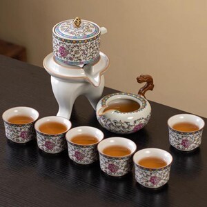 Op de afbeelding: Een keramische theeset met een witte standaard, een theepot, een serveerpot met een drakenhandvat en zes theekopjes. De set heeft bloemmotieven in roze, blauw en groen. De kopjes zijn gevuld met thee.