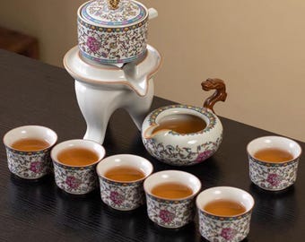 May include: A ceramic tea set with a white stand, a teapot, a serving pot with a dragon handle, and six teacups. The set features floral patterns in pink, blue, and green. The teacups are filled with tea.