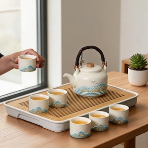 Puede incluir: Un juego de té de cerámica que incluye una tetera con asa envuelta en color oscuro y cinco tazas a juego. Cada pieza es de color blanco roto con un diseño en relieve de montañas azules y bronceadas y dos pequeños pájaros negros.