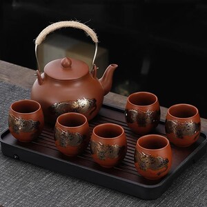Puede incluir: Un juego de té de cerámica marrón con una tetera y seis tazas a juego. La tetera tiene un asa tejida y las tazas y la tetera están decoradas con un diseño de dragón dorado. El juego se exhibe en una bandeja negra.