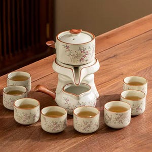 May include: A ceramic tea set with a teapot, infuser, and multiple teacups. The set is white with floral designs in pink and green. The teapot has a brown handle and lid. The teacups are filled with tea and arranged on a wooden surface.