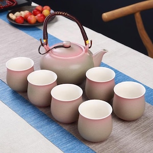 Peut inclure: Service à thé en céramique avec une théière dégradée rose et verte, une anse tressée et six tasses assorties. Les tasses et la théière ont une finition lisse et mate. L'ensemble est disposé sur une nappe à motifs.