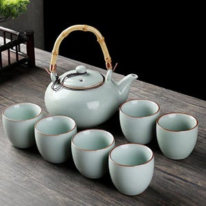 Op de afbeelding: Een celadongroene keramische theeset met een theepot en zes bijpassende kopjes. De theepot heeft een bamboe handvat en een deksel. De kopjes hebben een gouden rand. De set staat op een houten oppervlak.