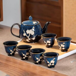 May include: A ceramic tea set featuring a navy blue teapot and six matching cups. The set has a floral design in white and gold accents. The teapot has a black handle and spout. The cups are small and round.