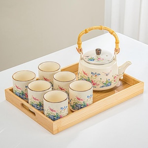 Può includere: Un set da tè con una teiera con manico in bambù e sei tazze abbinate, il tutto su un vassoio di legno. La teiera e le tazze sono decorate con motivi floreali e di uccelli nei toni del rosa, blu e verde.