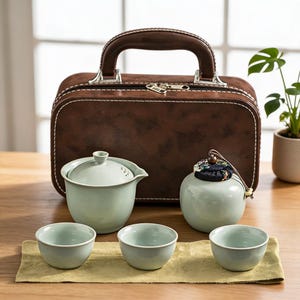 Könnte beinhalten: Ein Teeservice bestehend aus einer blassgrünen Keramik-Teekanne mit Deckel, einer passenden Teedose mit dunklem Stoffdeckel mit Blumenmuster und drei kleinen Teetassen. Das Set steht vor einem braunen, rechteckigen Transportkoffer.