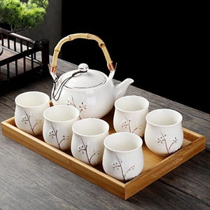 Peut inclure: Service à thé en céramique blanche comprenant une théière et six tasses assorties, toutes décorées d'un motif floral délicat. La théière a une poignée en bambou et repose sur un plateau en bois. Les tasses sont disposées autour de la théière.