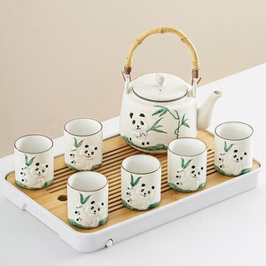 Puede incluir: Juego de té de cerámica blanca con una tetera y seis tazas, cada una adornada con motivos de osos panda y hojas de bambú. La tetera tiene un asa de bambú. El juego se presenta sobre una bandeja de bambú. Medidas en cm.