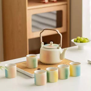Peut inclure: Un service à thé aux couleurs pastel comprenant une théière avec une poignée en bois et six tasses assorties. La théière et les tasses présentent un dégradé de couleurs, dont le rose, le jaune, le vert et le bleu. L'ensemble est disposé sur un plateau en bois.