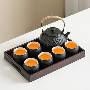 Puede incluir: Un juego de té negro sobre una bandeja de madera. El juego incluye una tetera negra texturizada con un asa tejida, seis tazas de té negras con interiores blancos y una bandeja llena de líquido naranja. El juego de té está dispuesto sobre una superficie blanca.