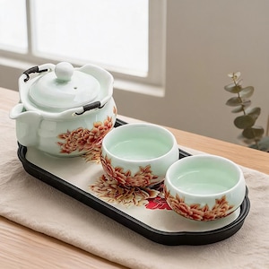 Könnte beinhalten: Ein Keramik-Teeservice bestehend aus einer hellgrünen Teekanne mit Deckel und zwei passenden Teetassen, die alle mit orangefarbenen und roten Blumenmustern verziert sind. Das Set steht auf einem ovalen schwarzen Tablett mit cremefarbenem Boden.